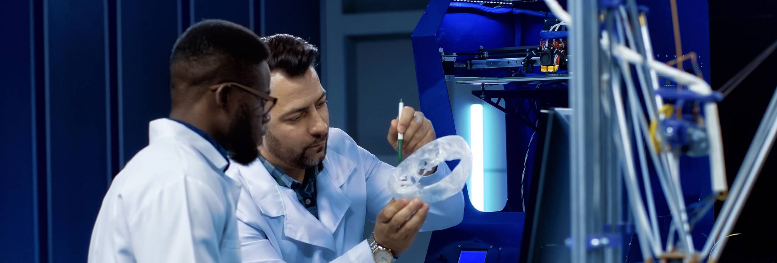 Two men in lab coats work together, one holding a transparent ring while the other uses a tool, with a 3D printer in the background.